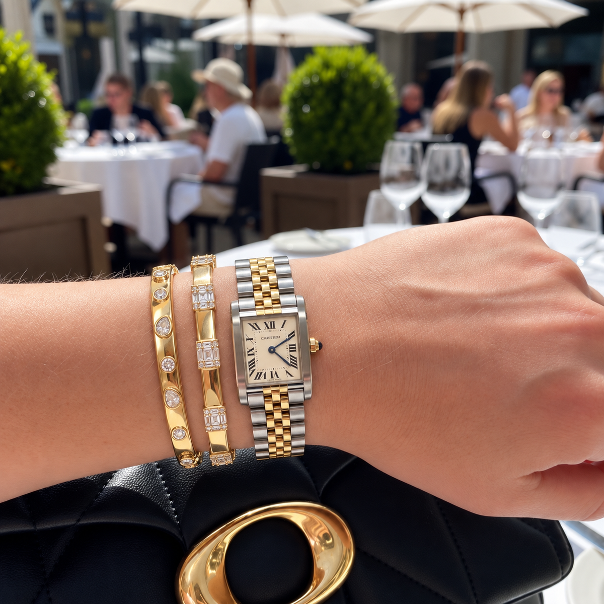 A close-up of a person's wrist wearing a two-tone stainless steel and  gold watch with a diamond-embellished bangle bracelets,  set against a blurred outdoor dining background.