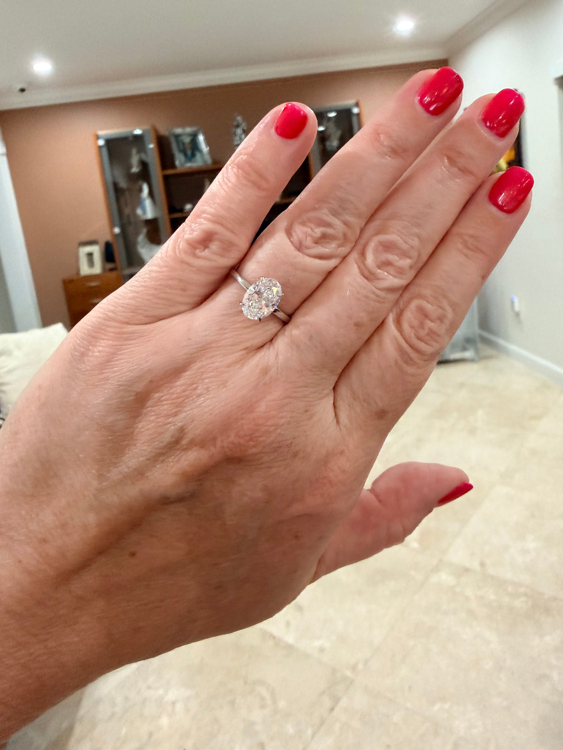 A close-up of a hand wearing a white gold engagement ring with an oval-cut diamond gemstone, set against an indoor home background.