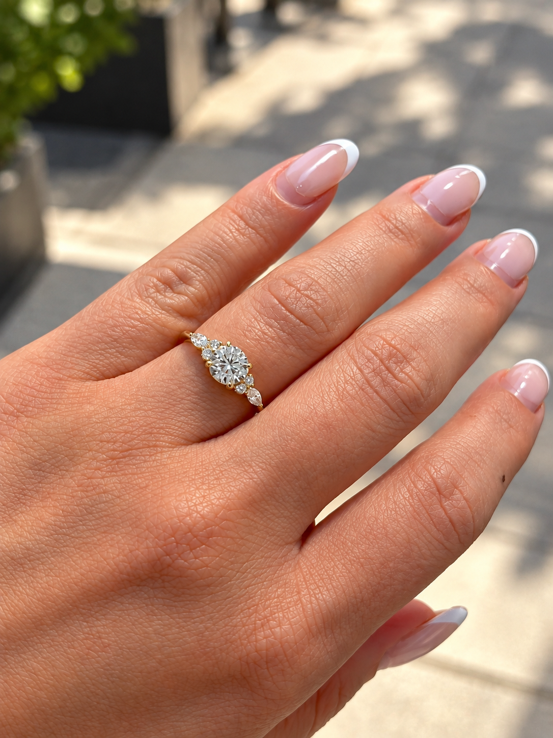 A close-up of a hand with a French manicure wearing a yellow gold  engagement diamond ring with a marquise-shaped cluster of  side diamond stones.