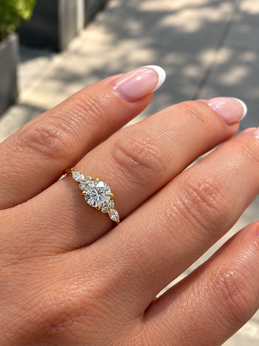 A close-up of a hand wearing a yellow gold engagement ring with a round brilliant cut diamond on the center and  marquise cut diamonds and smaller round diamonds on either side, set against an outdoor blurred background. 