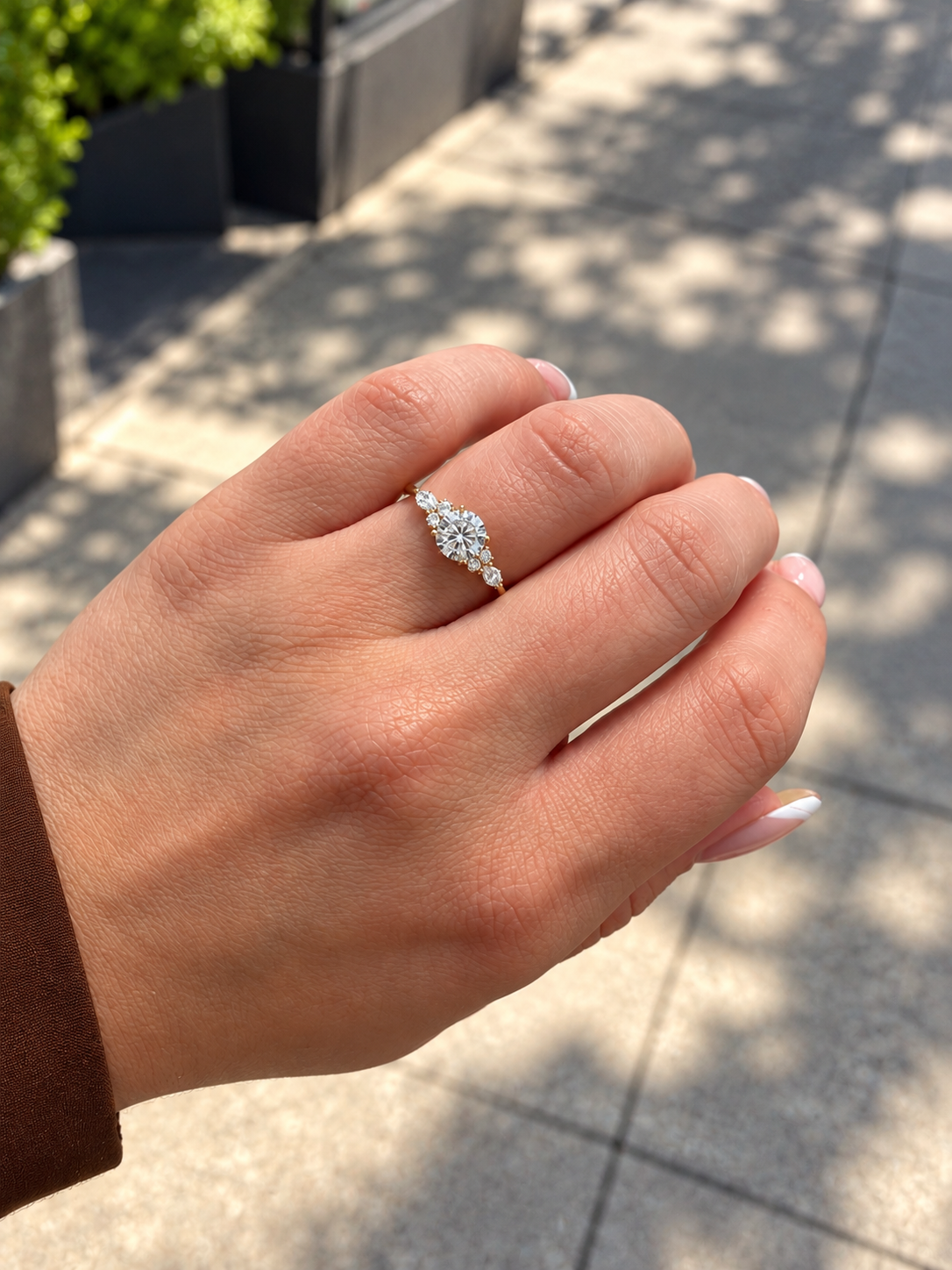 A close-up of a hand wearing a yellow gold engagement ring with a round brilliant cut diamond on the center and  marquise cut diamonds and smaller round diamonds on either side, set against an outdoor blurred background. 