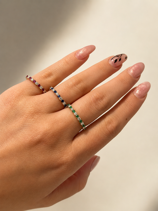 A close-up of a hand wearing three delicate gold band rings with small colorful  emerald, ruby, blue sapphire and diamond gemstones, set against a soft beige background.