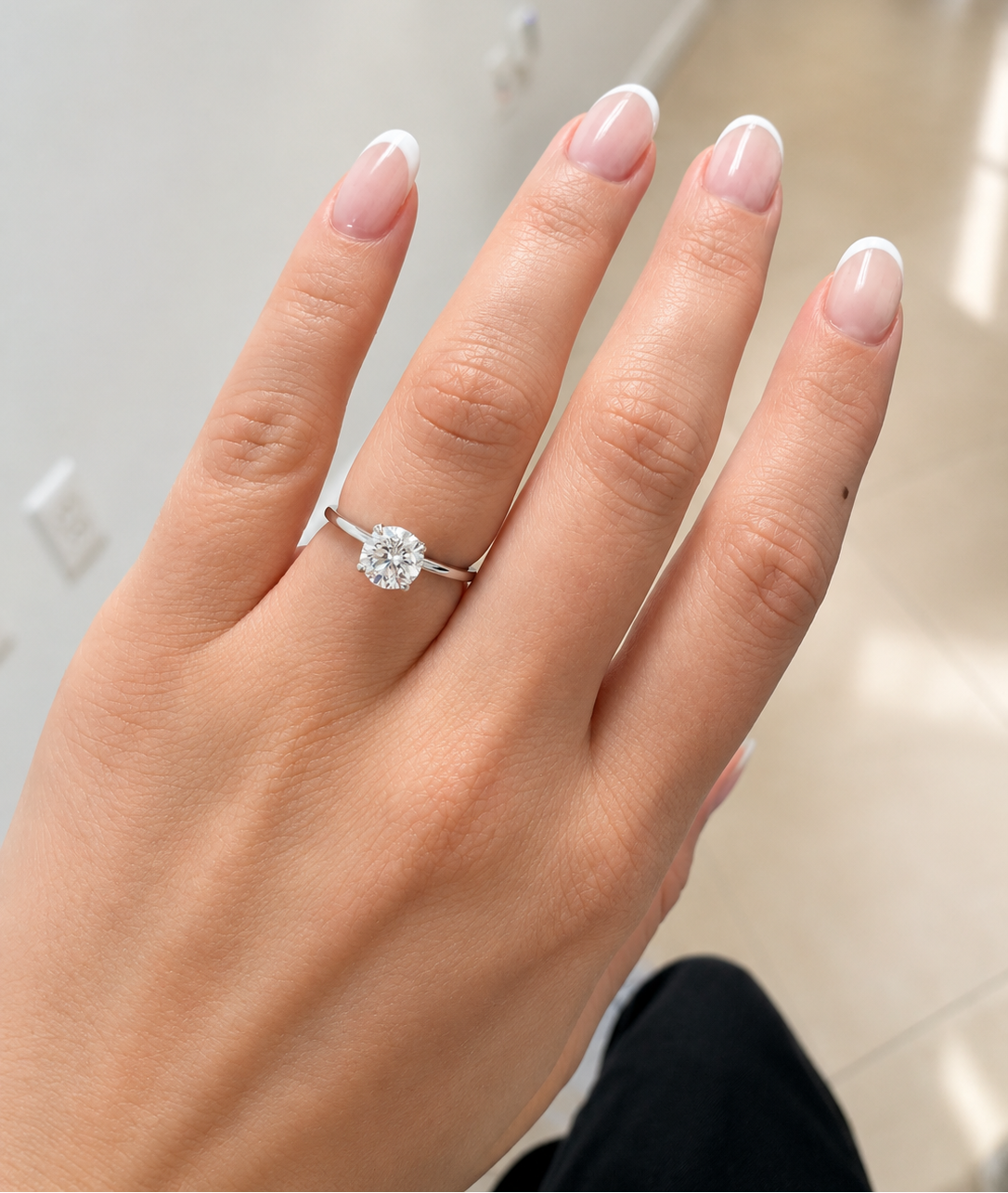A close-up of a hand wearing a classic solitaire engagement ring with a round brilliant-cut diamond on a thin silver band, set against an indoor background.