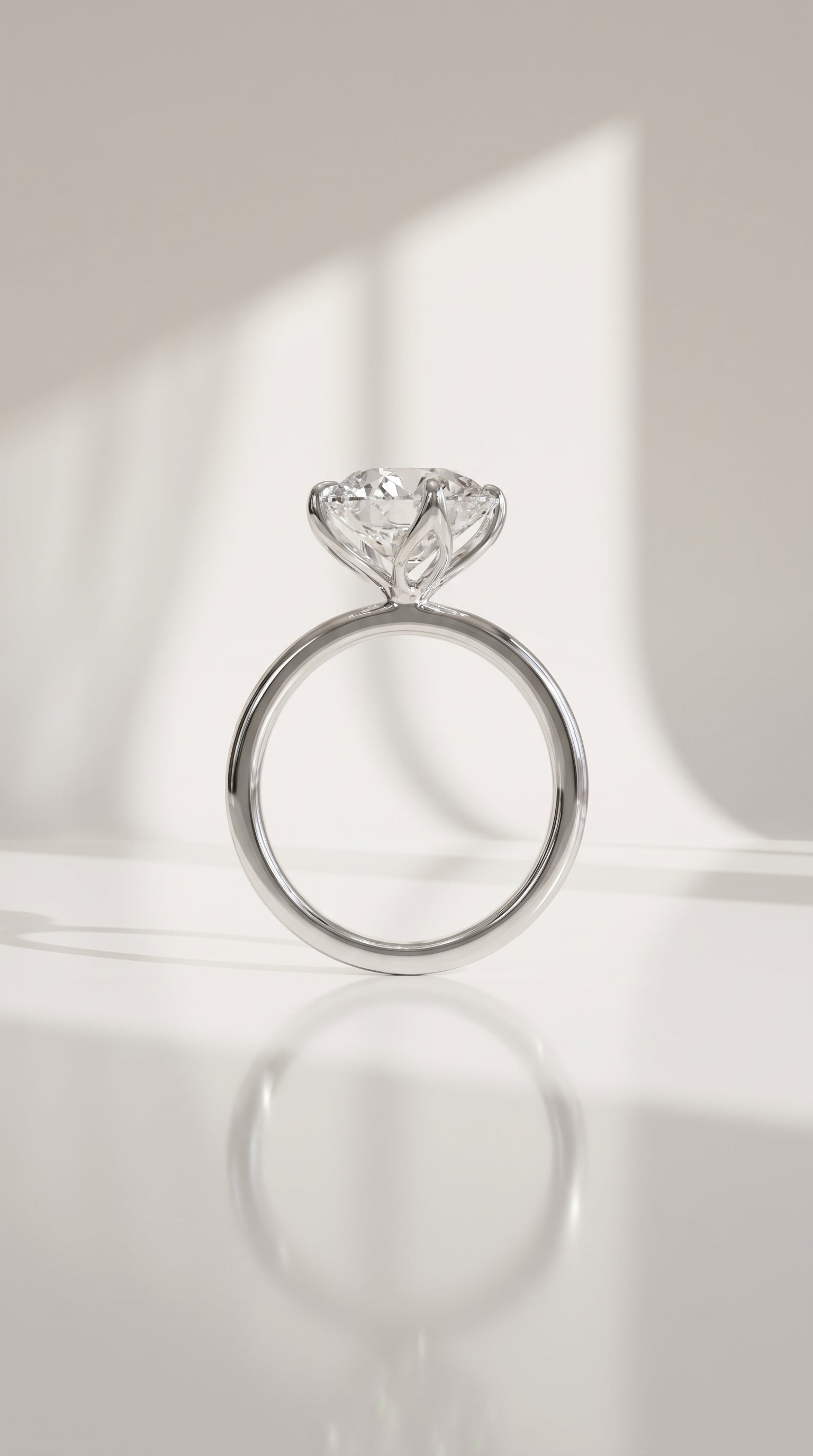 A white gold solitaire engagement ring with a round-shaped diamond gemstone, displayed on a reflective white surface with soft window light shadows.
