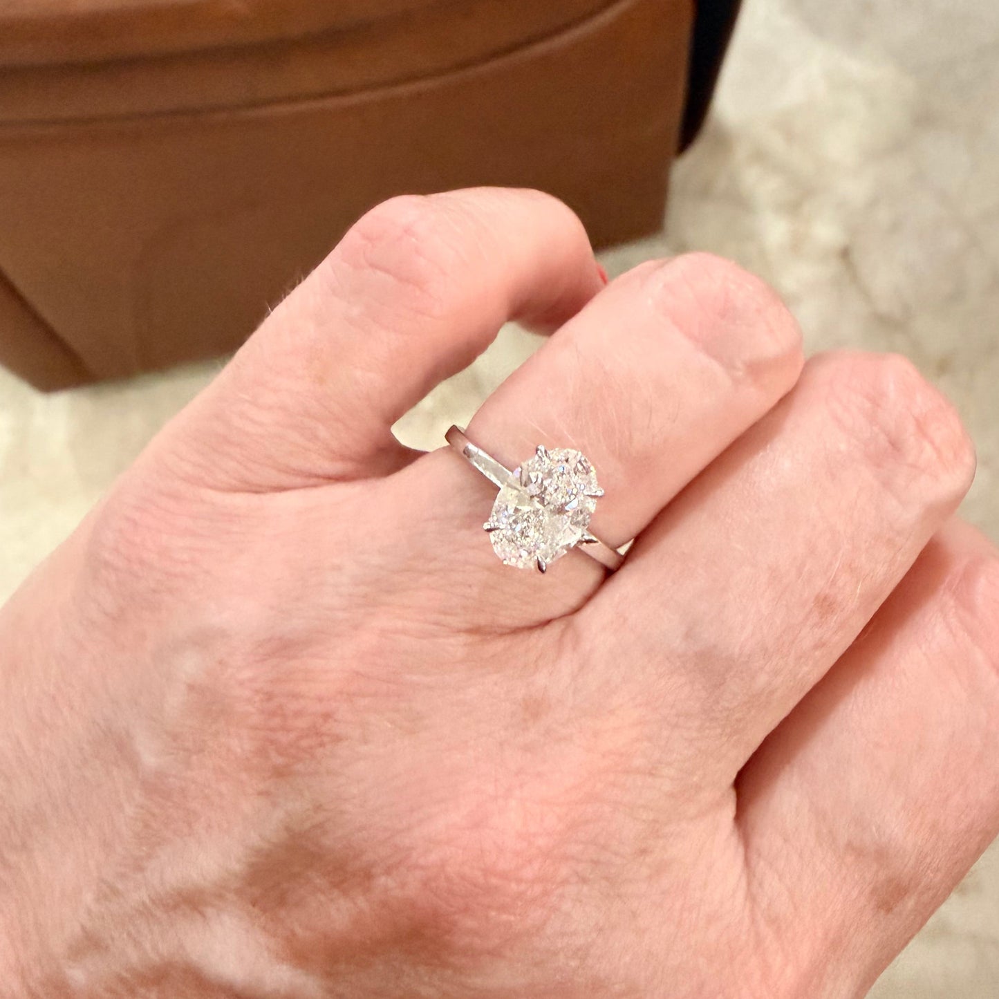 A close-up of a hand wearing a white gold engagement ring with a large, oval cut, diamond gemstone in a solitaire setting, photographed in an indoor setting with a brown leather chair in the background.