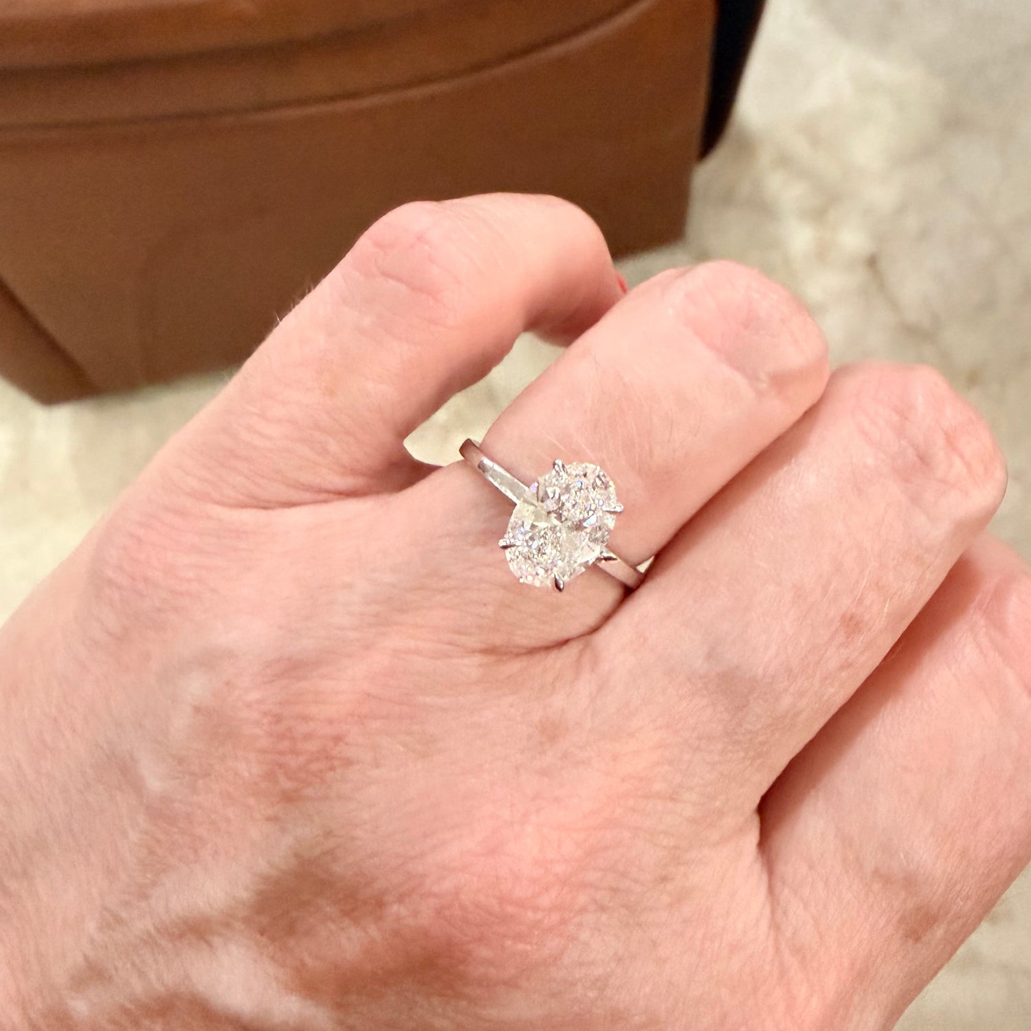 A close-up of a hand wearing a white gold engagement ring with a large, oval cut, diamond gemstone in a solitaire setting, photographed in an indoor setting with a brown leather chair in the background.