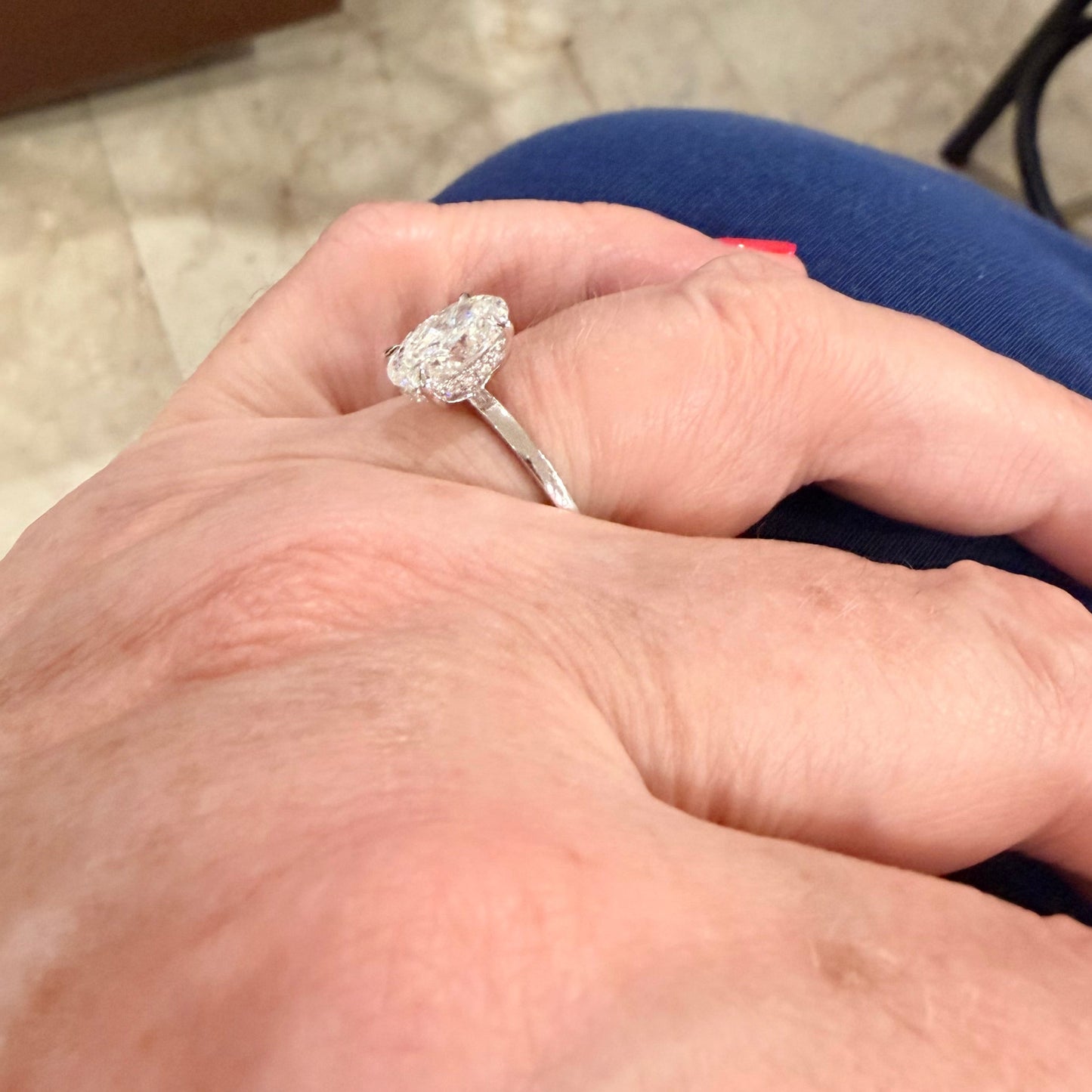 A close-up of a hand wearing a white gold engagement ring with a oval-cut diamond gemstone, resting on blue fabric in an indoor setting.