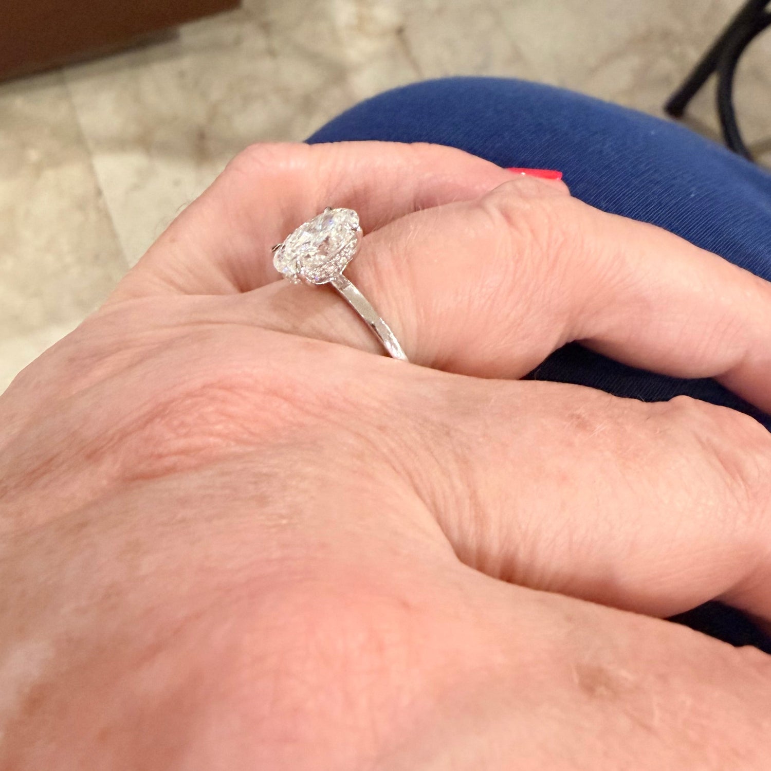 A close-up of a hand wearing a white gold engagement ring with a oval-cut diamond gemstone, resting on blue fabric in an indoor setting.