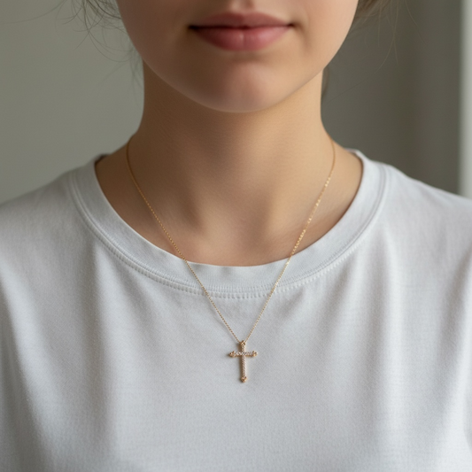A close-up of a person wearing a delicate gold cross pendant necklace on a fine chain, paired with a white crew-neck t-shirt.