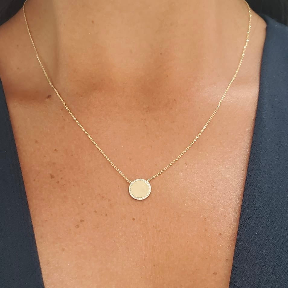 A delicate gold chain necklace with a small, flat circular disc pendant, shown worn on a model's neck against a dark navy blue top.