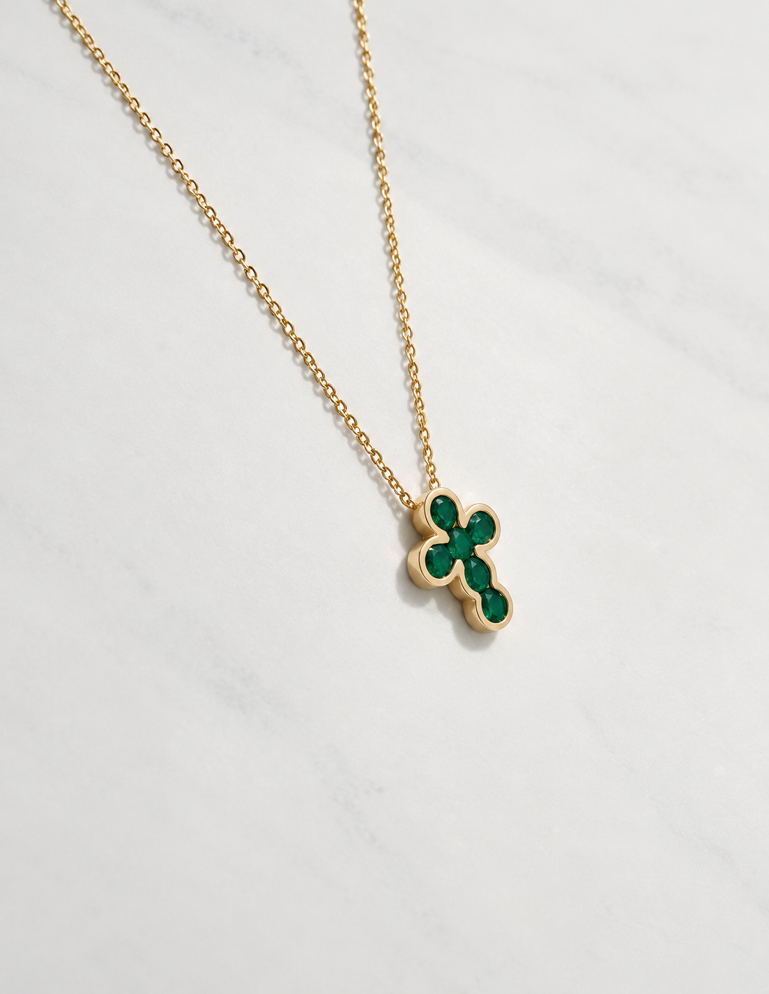 A gold chain necklace with cross pendant featuring inlaid green emerald stones, displayed on a light gray marble surface.