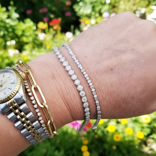 A close-up of a person's wrist wearing multiple bracelets, including a silver tennis bracelet, a silver beaded bracelet, a gold paperclip chain bracelet, and a gold beaded chain bracelet, alongside a gold and silver watch. The background is a blurred green garden with colorful flowers.