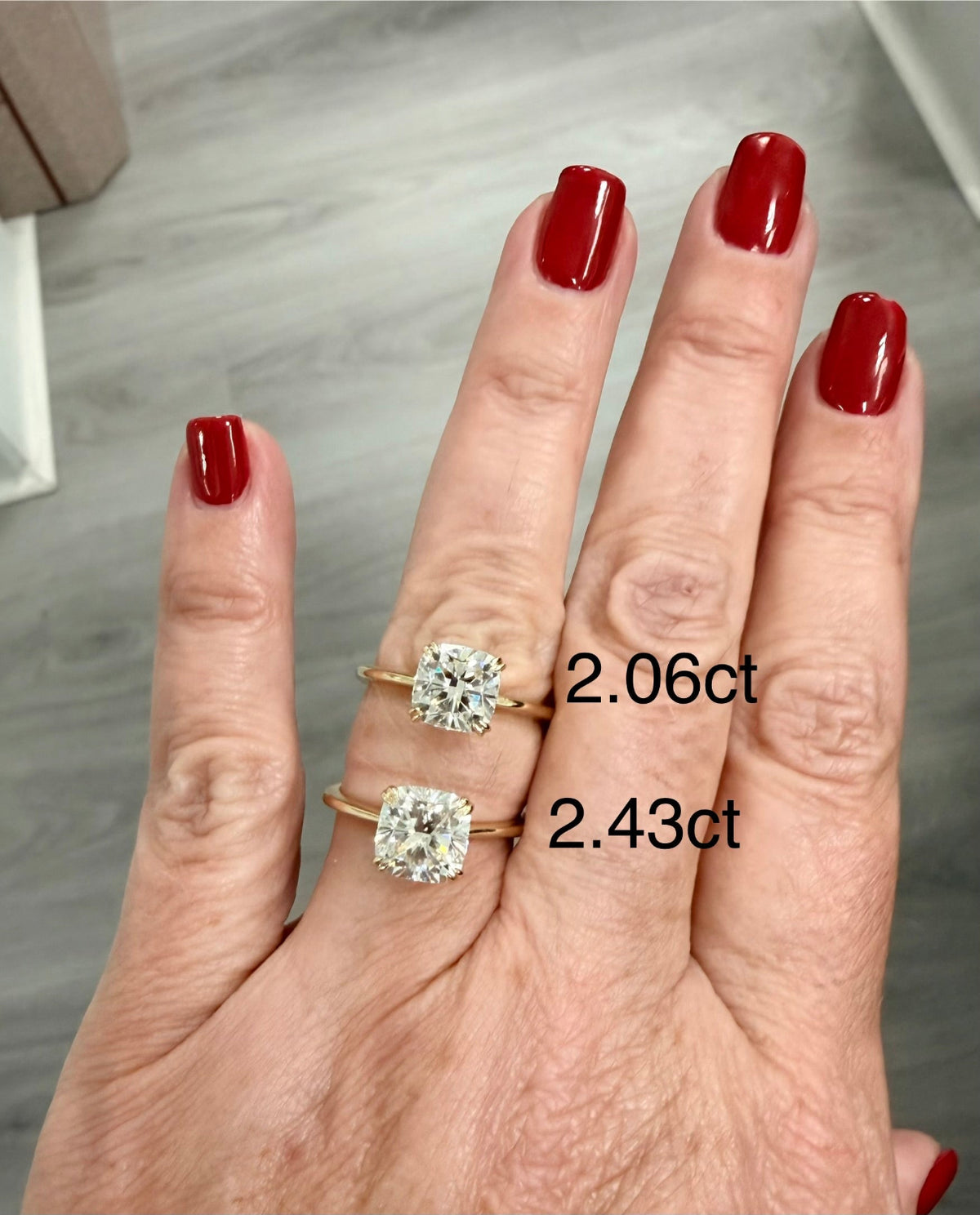 A close-up of a hand with red nail polish wearing two gold rings with cushion-cut lab diamond gemstones, one labeled 2.06ct and the other 2.