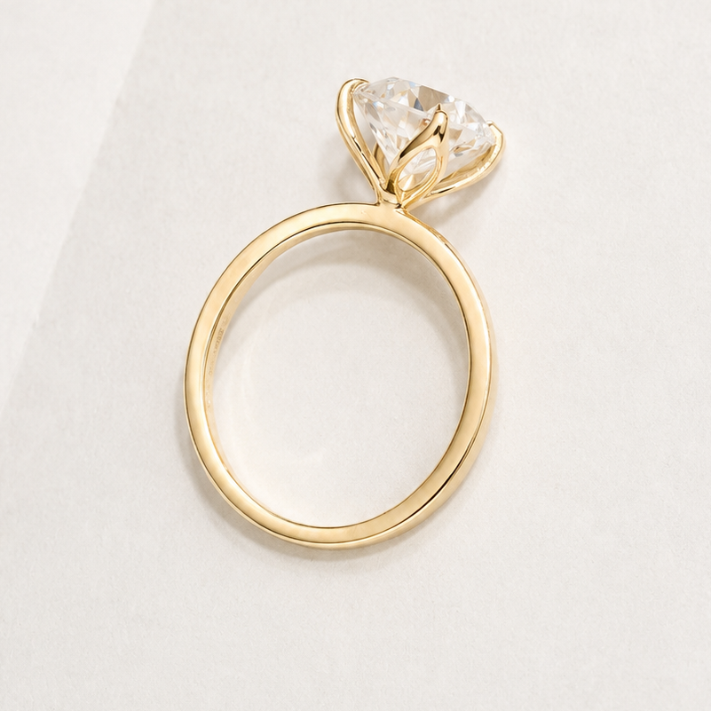 A yellow gold engagement ring with a round-shaped diamond gemstone in a prong setting, displayed on a clean, light off-white background.
