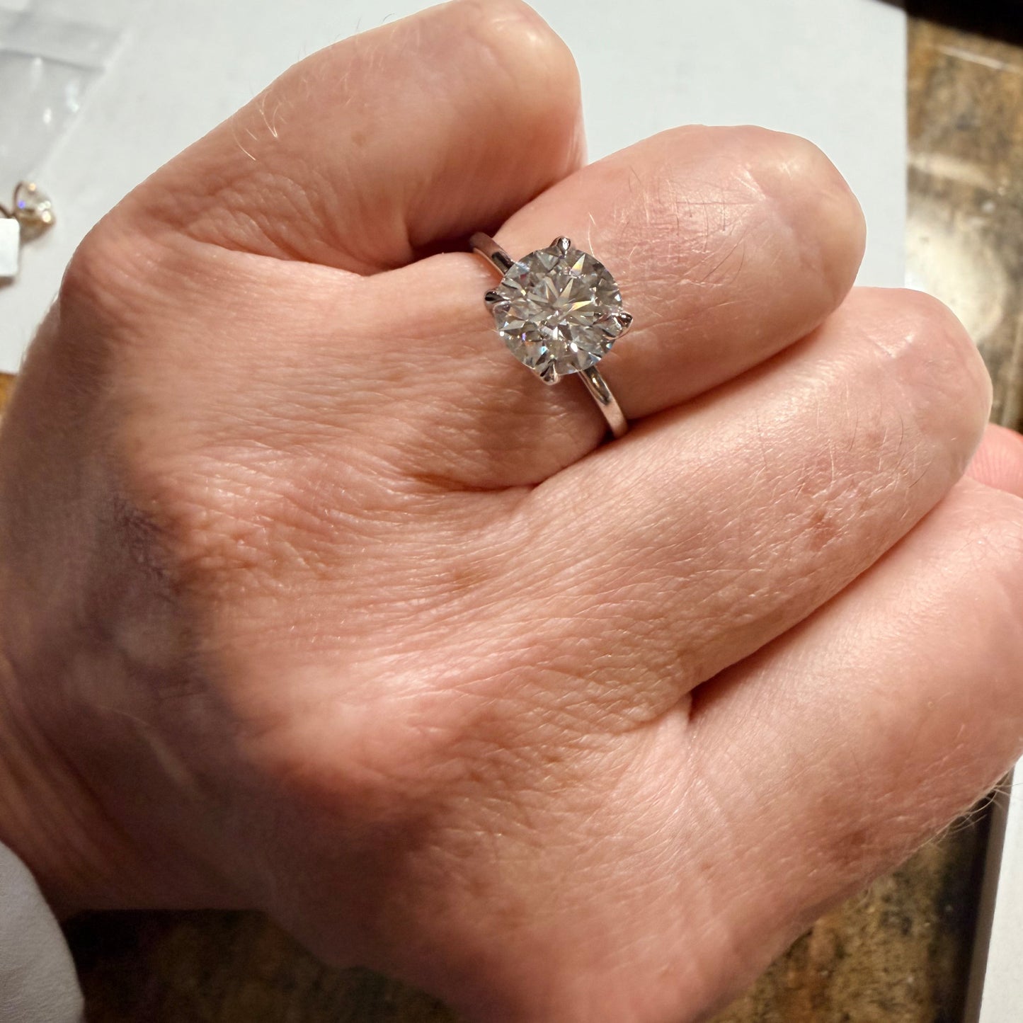 A close-up of a hand wearing a white gold engagement ring with a round brilliant-cut diamond gemstone, set against a casual tabletop background.