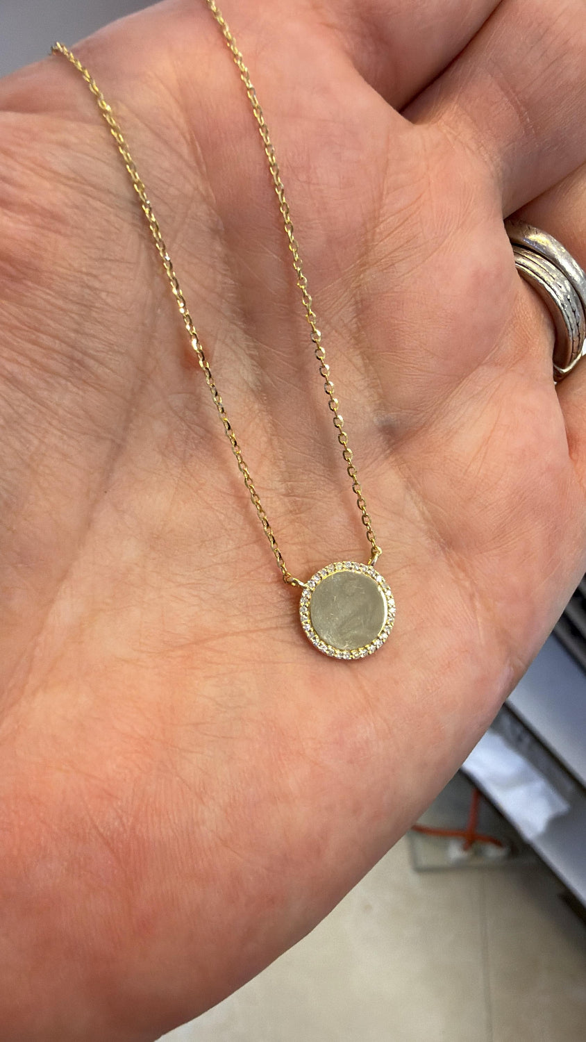 A gold chain necklace with a round high polish gold pendant set with small diamond pave stones, held in a person's hand.