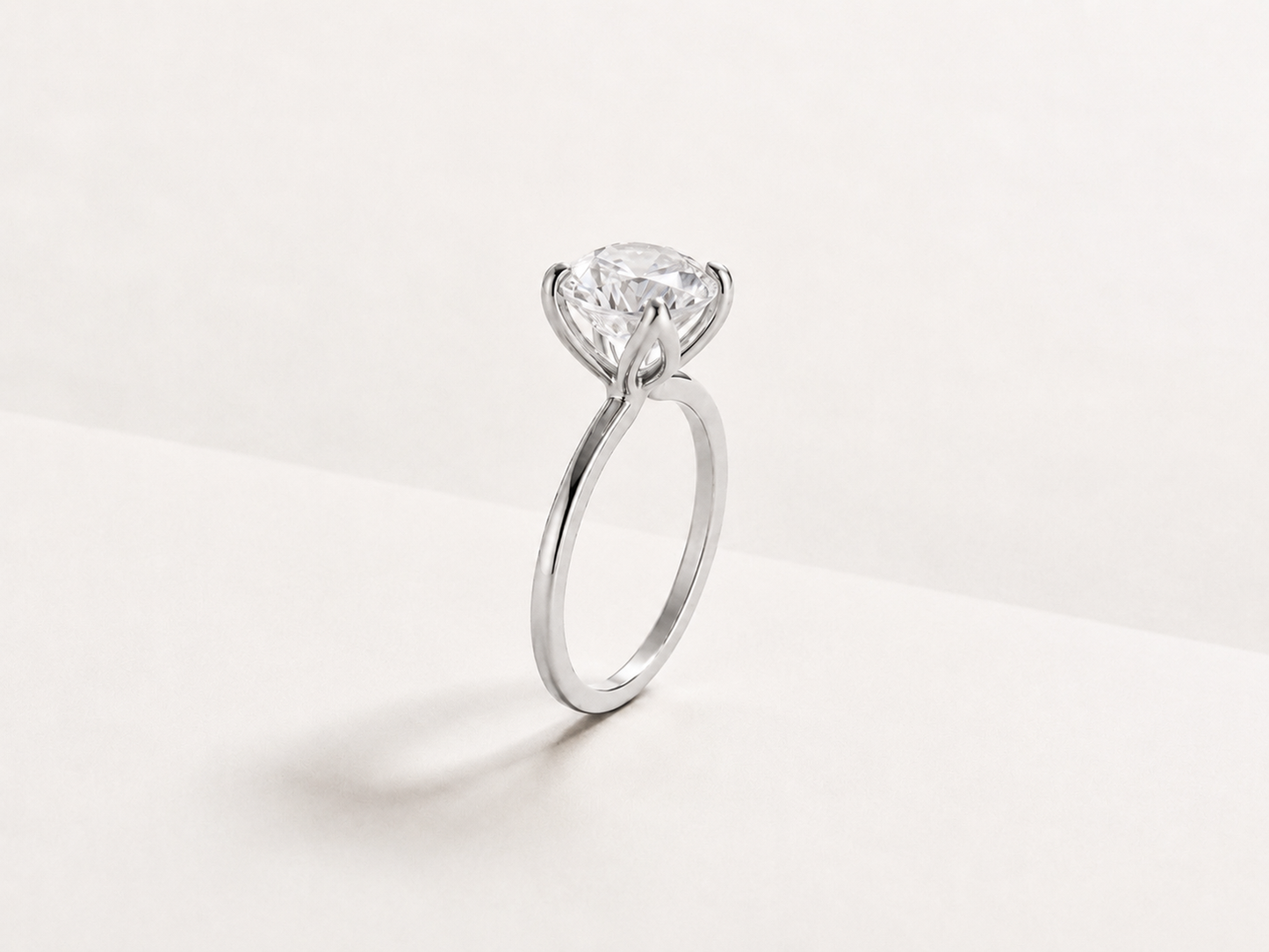 A classic solitaire engagement ring featuring a round brilliant-cut diamond set in a white gold metal band, photographed on a clean, light off-white background.