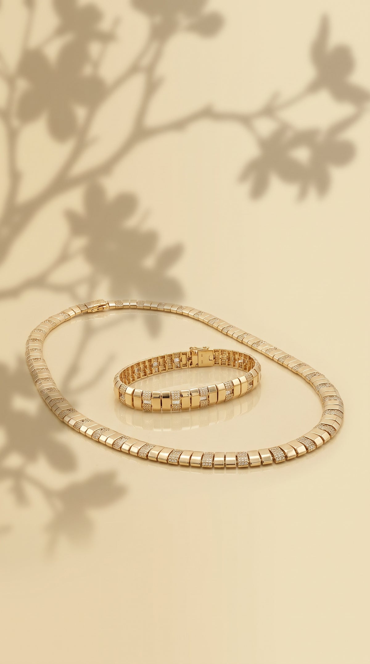 A gold wide contemporary diamond tennis bracelet and matching linked necklace displayed on a warm beige background with soft leaf shadows.