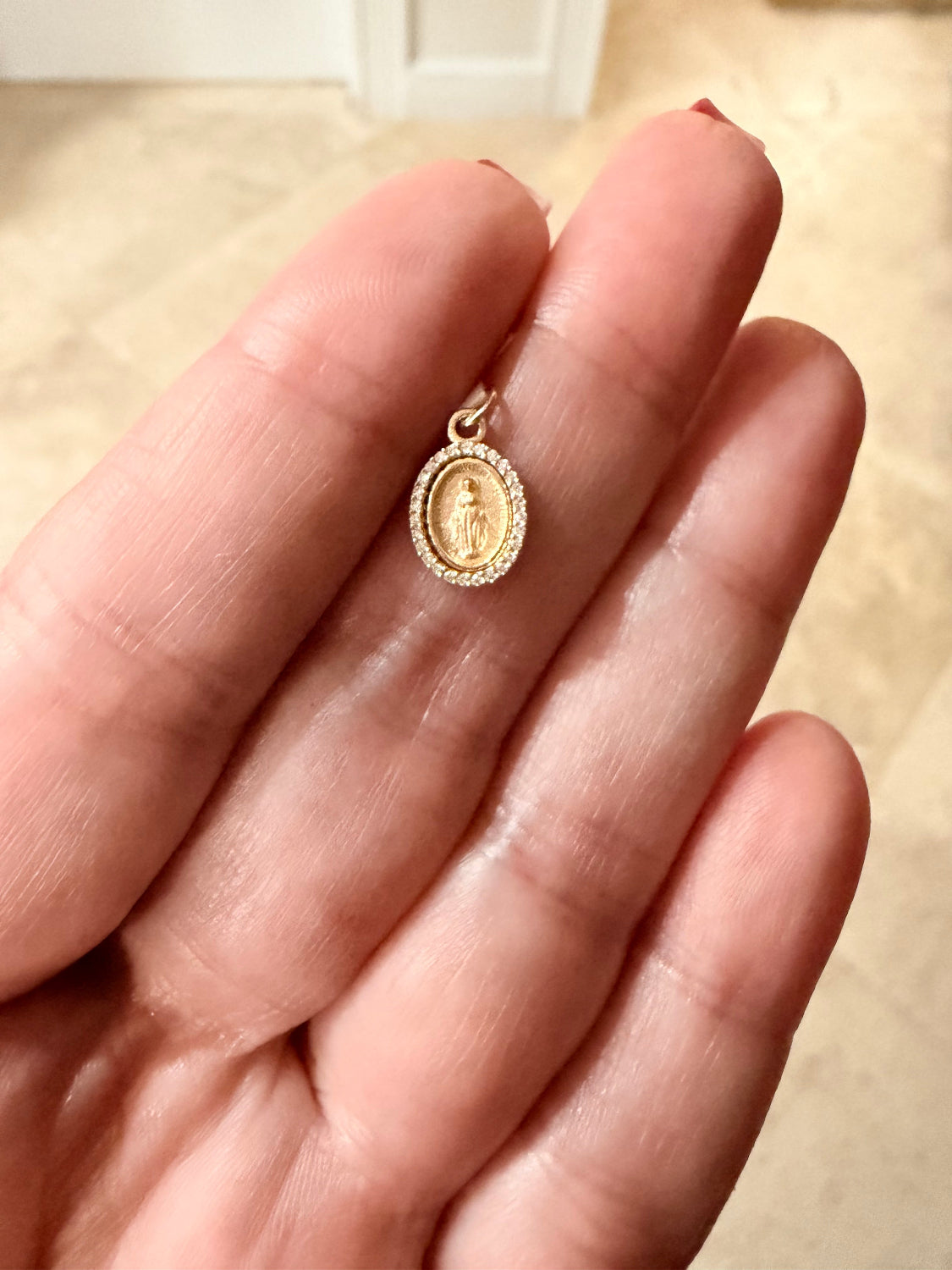 A small oval  Medal gold pendant with a diamond halo and central Miraculous Medal religious figure, resting on a person's fingers against a light beige tiled background.