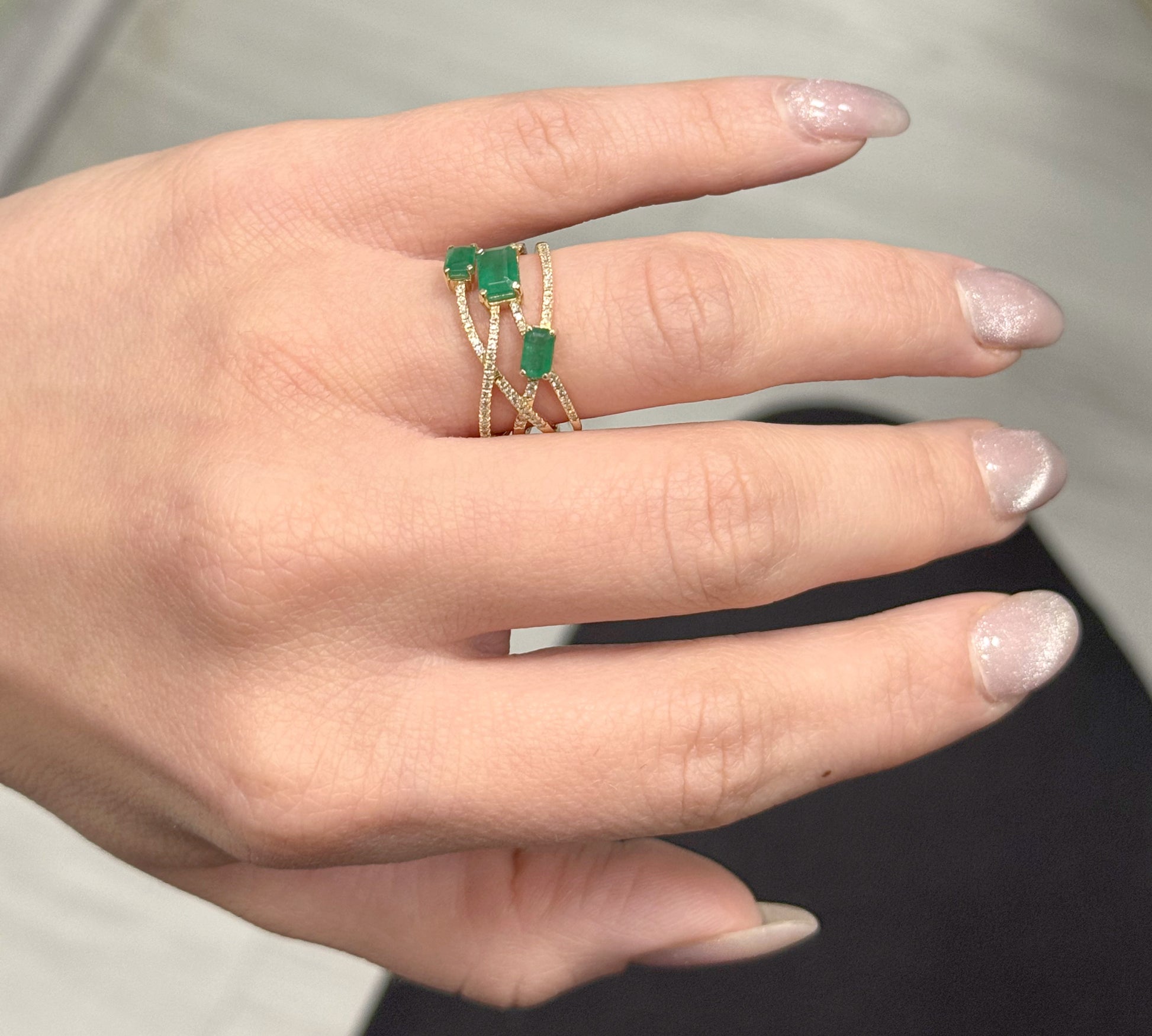 Hand wearing gold rings with emerald stones on a neutral background