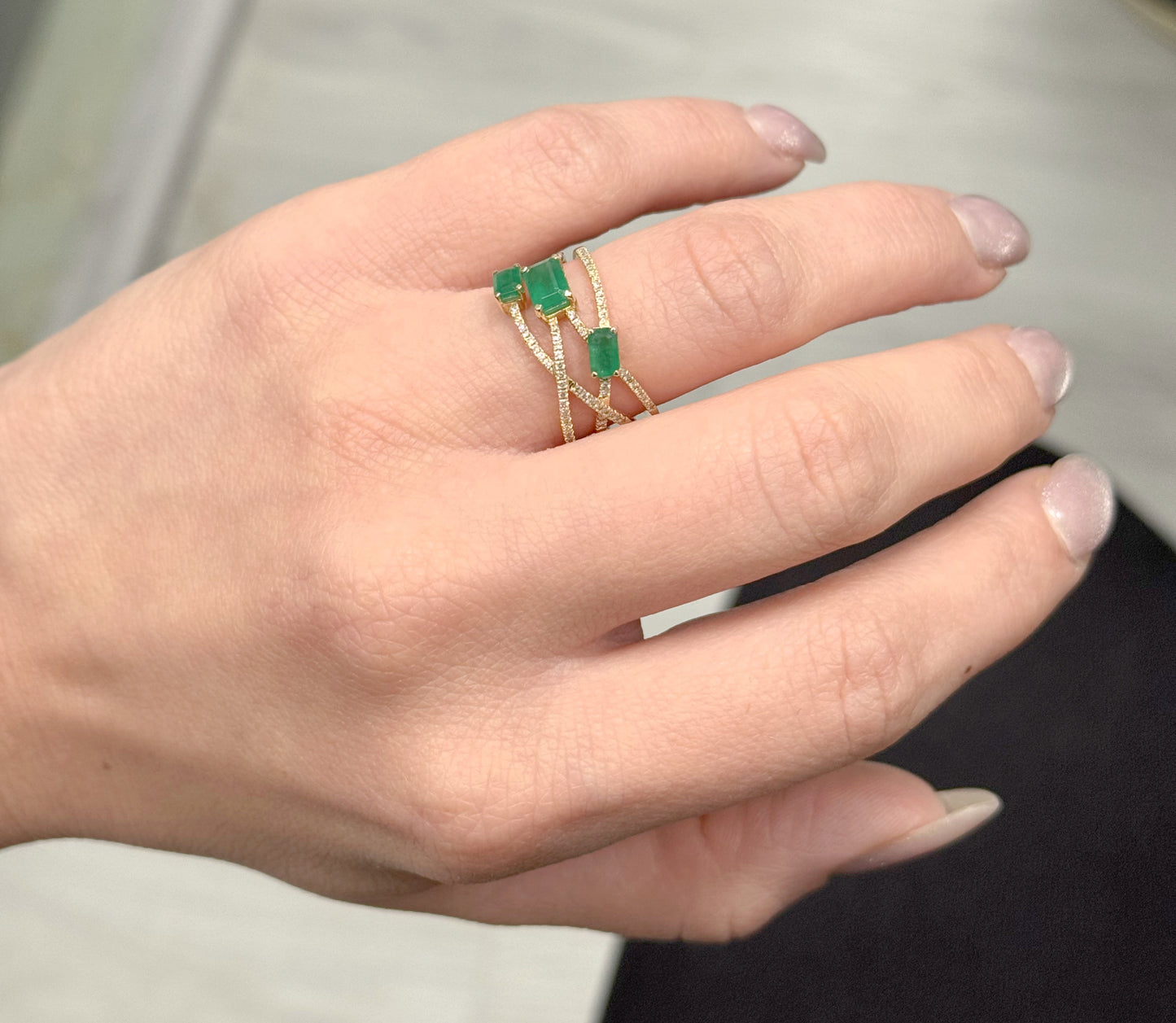Hand wearing a gold ring with green gemstones against a neutral background