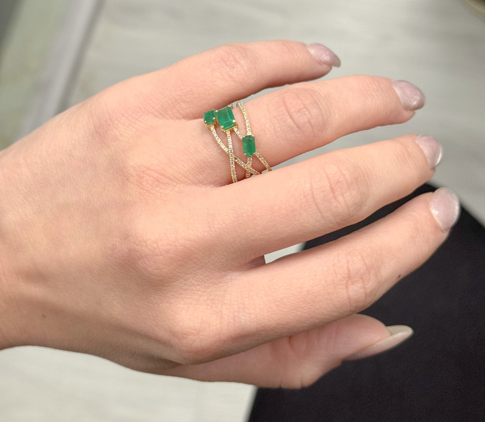 Hand wearing a gold ring with green gemstones against a neutral background