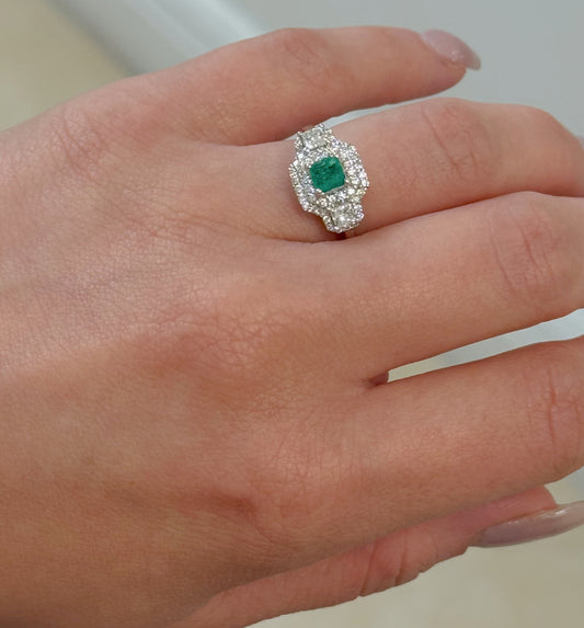 Hand wearing a ring with a green Emerald gemstone and diamond accents on a neutral background