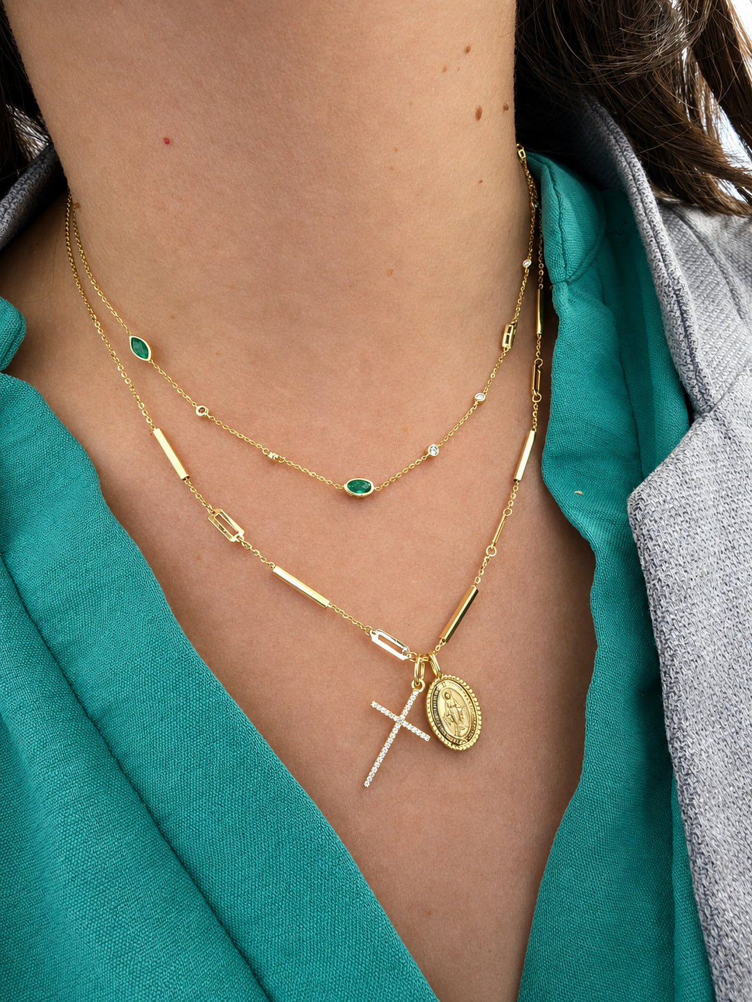 A close-up of a person's neck and chest wearing a layered gold necklace set. The set includes a delicate chain with small green oval stones, a chain with gold bar accents, and a longer chain featuring a diamond cross pendant, a gold oval Miraculous medallion.