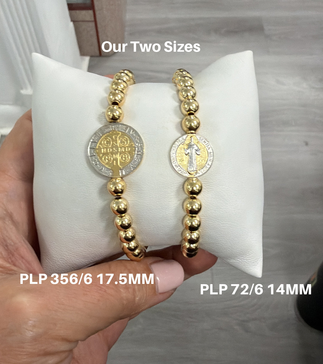 Two gold beaded bracelets with two sizes of the Saint Benedict medallions on a white cushion, held by a hand.