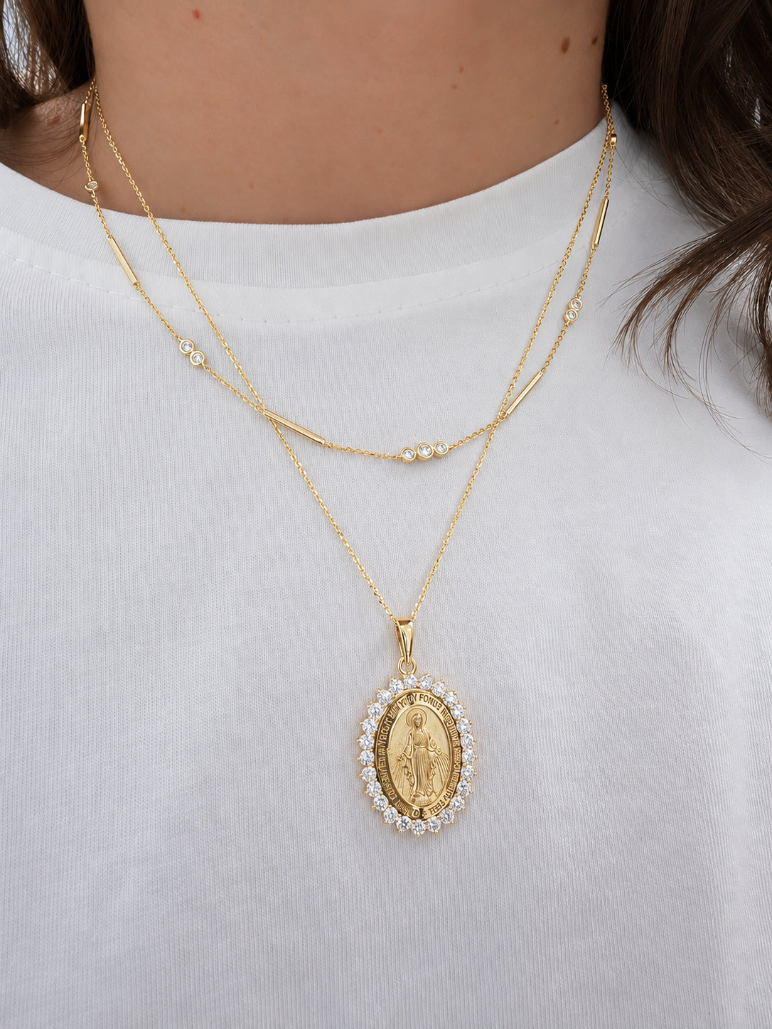 A close-up of a model wearing a double-layered gold necklace with a Miraculous Medal pendant. The necklace features one layer with a delicate chain and small diamond bezels, and the other layer with a thicker bar, both culminating in an oval gold Miraculous medal  pendant surrounded by a halo of small diamonds.