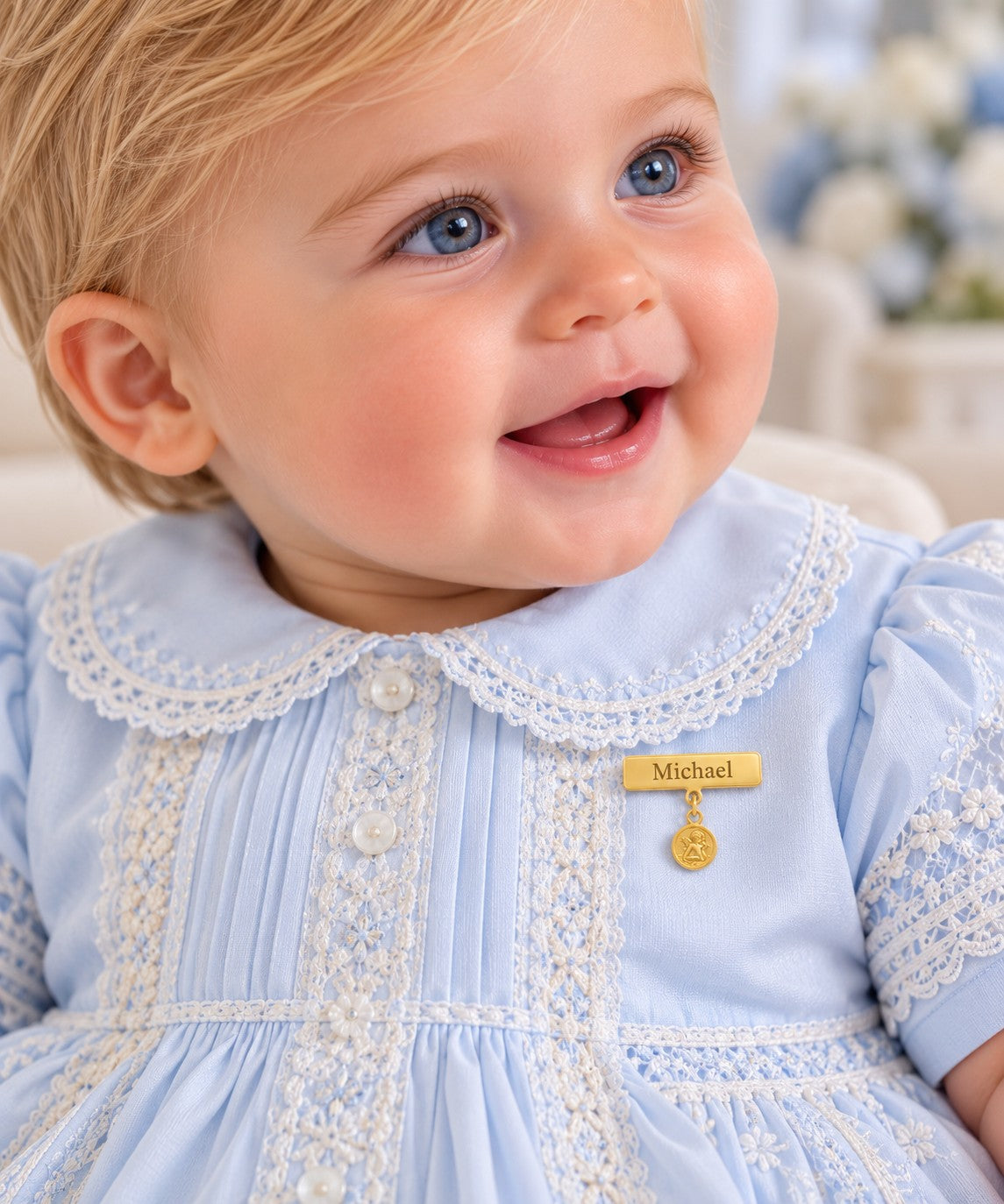 A close-up of a smiling baby wearing a light blue gown with white lace trim and an engravable gold plate baby pin with a hanging angel medal that reads 'Michael'.
