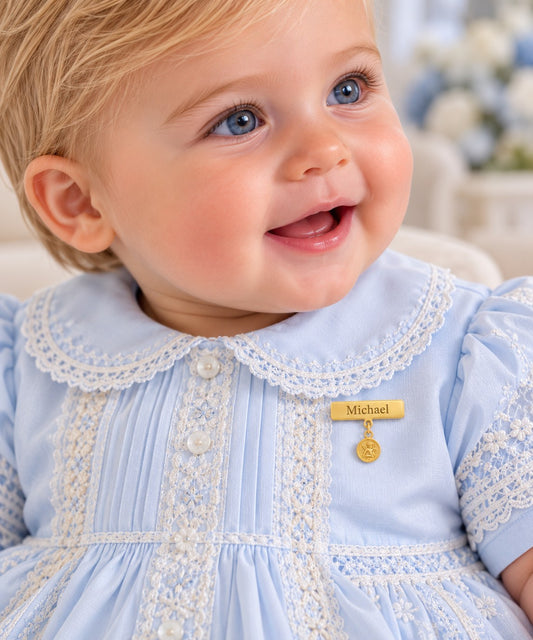 A close-up of a smiling baby wearing a light blue gown with white lace trim and an engravable gold plate baby pin with a hanging angel medal that reads 'Michael'.
