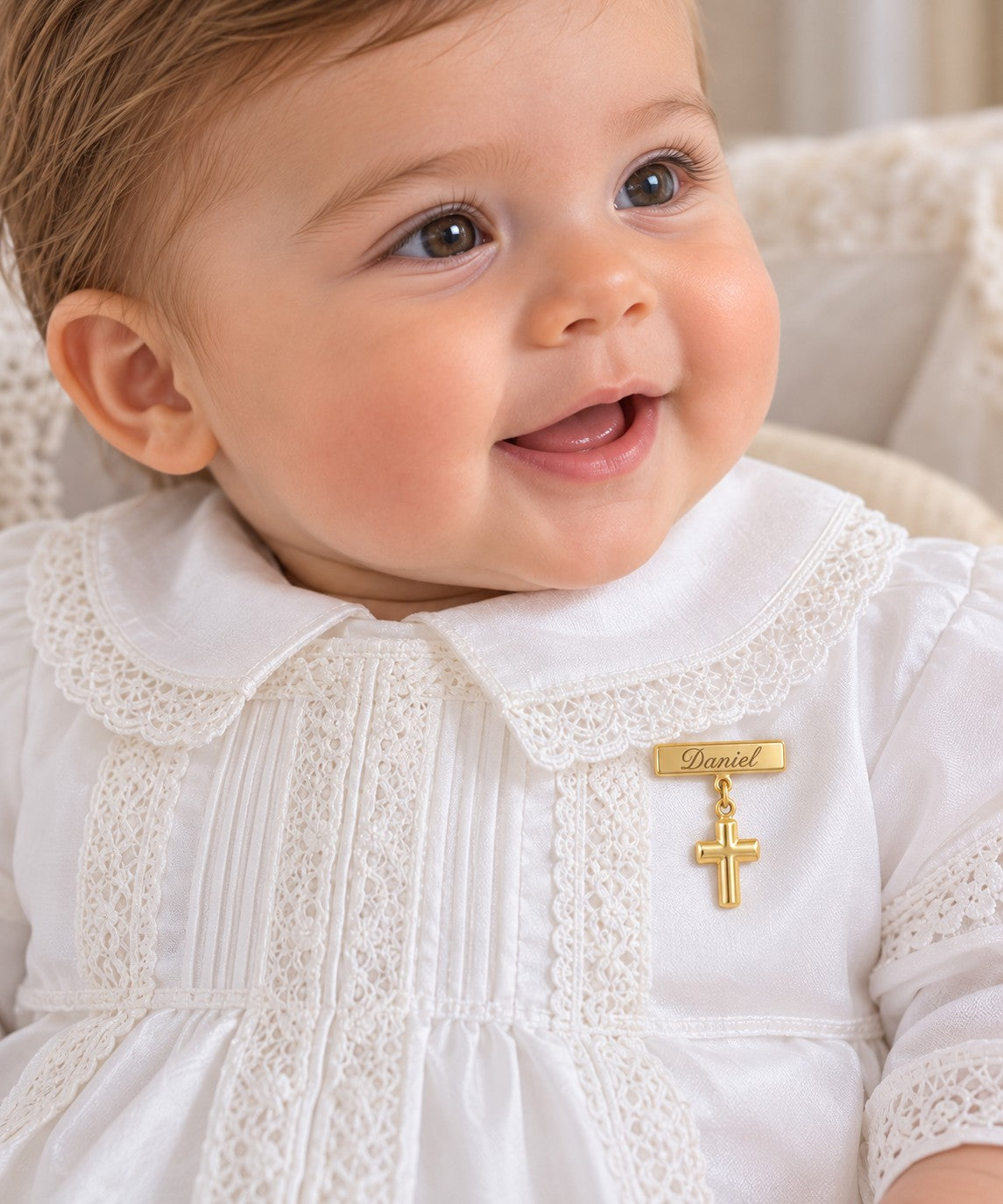 A close-up of a smiling baby wearing a white baby dress with lace trim and a gold cross pin engraved with the name 'Daniel'.