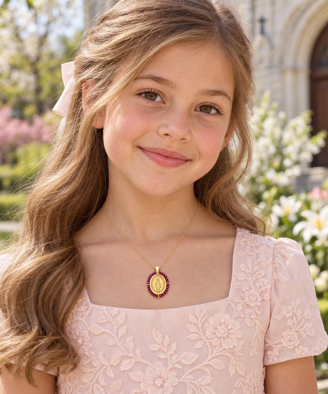 A young girl with long light brown hair wearing a pale pink floral dress and a gold pendant necklace with a red ruby beads around a Miraculous medallion, photographed outdoors in front of a blurred building and flowers.