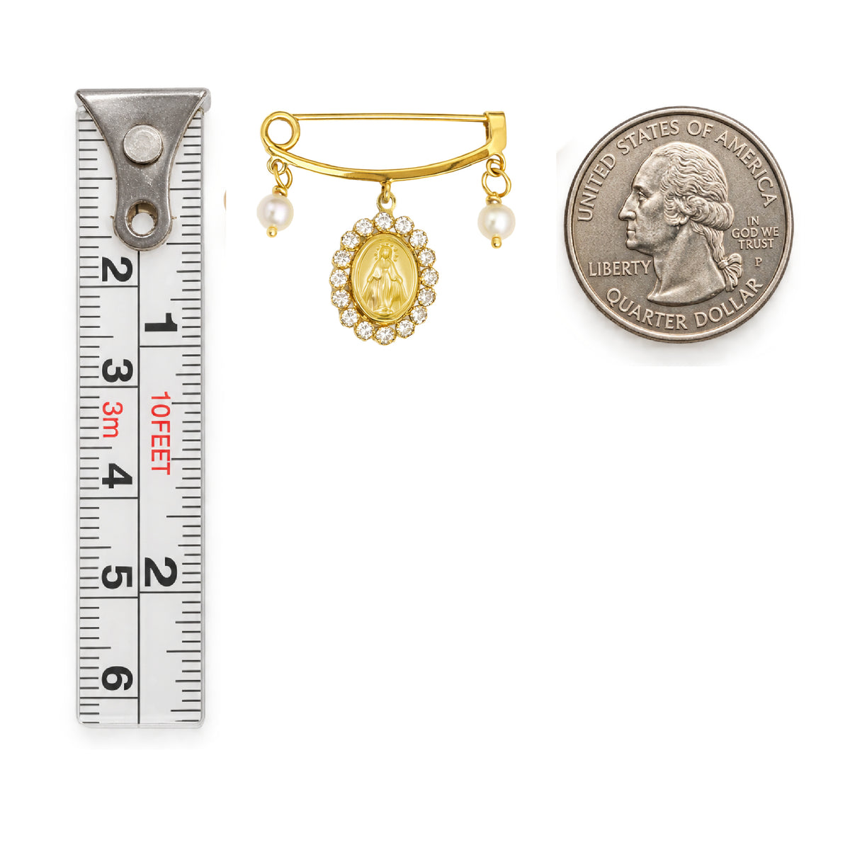 A gold Miraculous Medal brooch with pearl accents and a border of small clear stones, shown next to a metal ruler for scale and a US quarter coin for scale, on a plain white background.