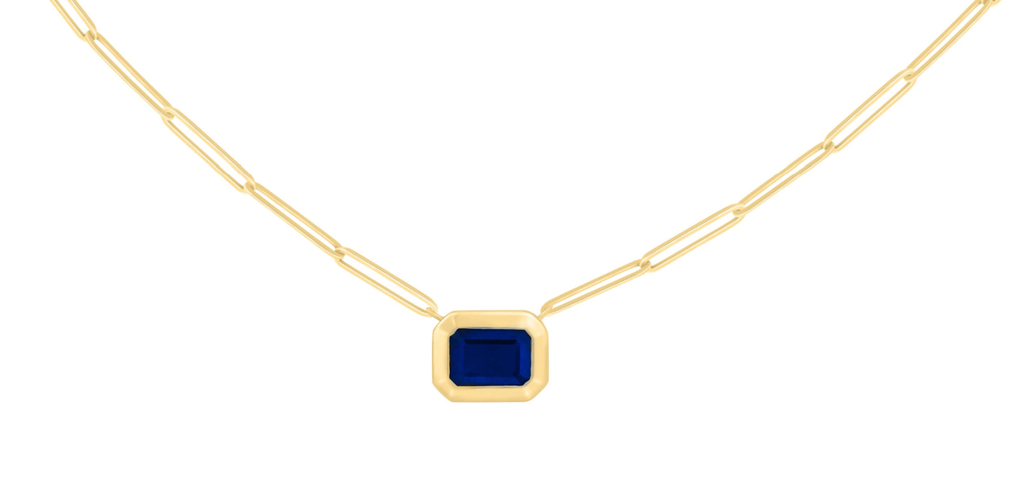 A 14K gold necklace featuring a bezel set step-cut Blue Sapphire color stone pendant on a paper clip chain.
