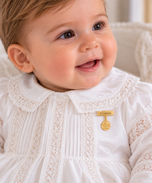 A close-up of a smiling baby wearing a white lace dress with a small gold angel medal pinned to the chest. The medal has a gold bar engraved with the name 'Cristian'.