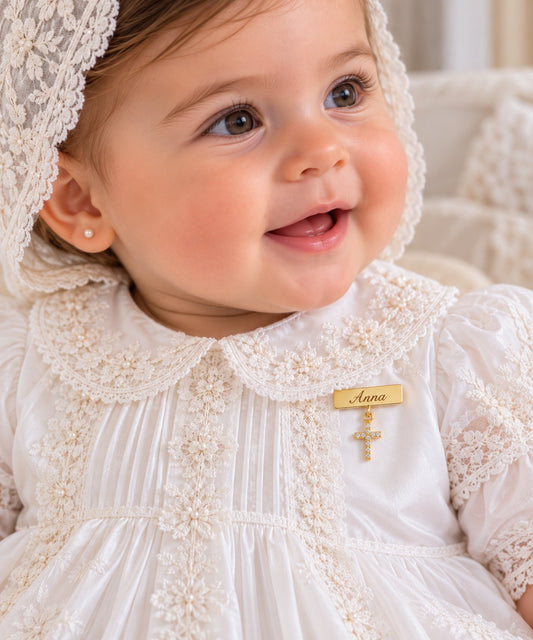 A close-up of a smiling baby wearing an ivory baptismal dress with lace trim, a matching lace bonnet, and a gold cross pin with a personalized name tag reading 'Anna'.