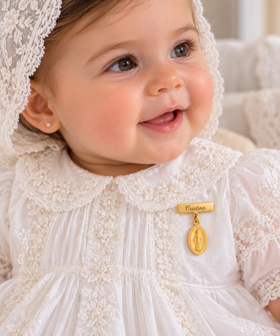 A close-up of a smiling baby wearing a white lace christening dress and bonnet, with a small gold Miraculous Medal pendant pinned to the chest on a small engravable plaque reading 'Cristina".