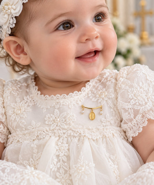 A close-up of a smiling baby wearing a white lace Baptismal Gown dress with a small gold Miraculous Medal baby pin at the chest, along with a matching floral headband.