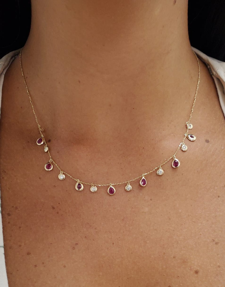 A delicate gold necklace with small teardrop-shaped red ruby gemstones and clear round diamond stones, shown worn on a person's neck and upper chest.