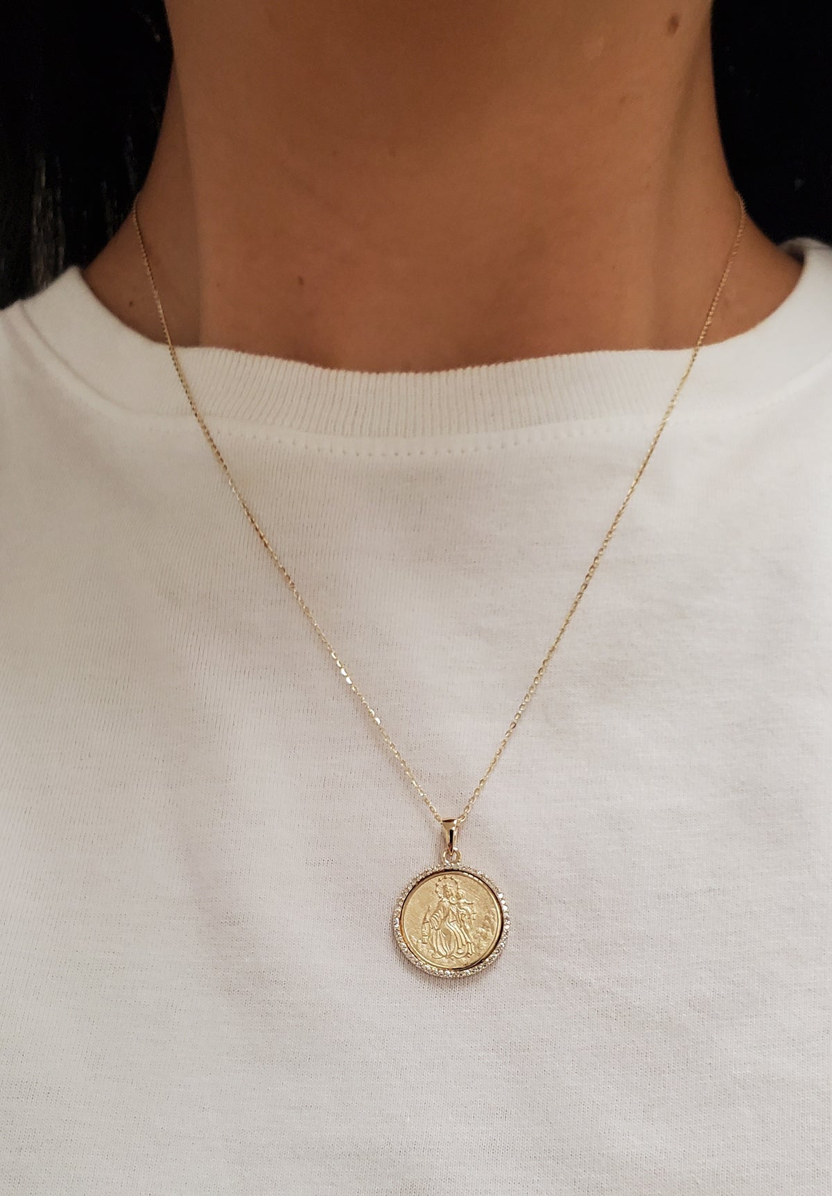 A 14K yellow gold Religious Medal pendant necklace worn by a person in a white crewneck shirt. The pendant features an embossed Our Lady of Mount Carmel (Virgen Del Carmen) design and is suspended from a delicate gold chain with a halo of natural diamonds.
