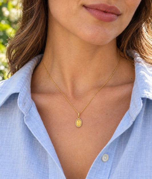 A close-up of a woman wearing a delicate gold chain necklace with an oval diamond Miraculous Medal pendant, paired with a light blue button-down shirt.